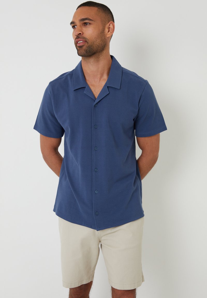 Korte mouwen overhemd met knopen in marineblauw, gemaakt van gladde stof, met een camp collar en vijf knopen aan de voorkant. Draagt met beige shorts.