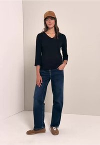 Schwarzes Langarmshirt, dunkelblaue Jeans, braune Mütze und braune Sneakers. Das Model steht mit einer Hand in der Tasche vor einem neutralen Hintergrund.