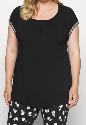 Kvinde iført en sort kortærmet top med stribet kant og sorte bukser med hvidt blomsterprint, stående mod en ensfarvet baggrund.