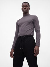 Ein Mann trägt ein eng anliegendes graues Langarmshirt und schwarze Hosen mit Kordelzug, steht vor einem einfarbigen weißen Hintergrund.