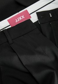 Zwarte broek met een gladde textuur, voorzien van een zichtbaar roze label met de tekst "JJXX" en een ritszak aan de zijkant.