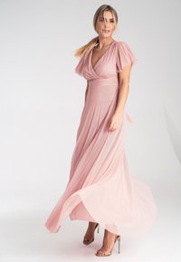 Rosa Chiffon-Maxi-Kleid mit kurzen, flatternden Ärmeln, Wickel-Oberteil und fließendem Rock. Das Kleid hat eine taillierte Taille und eine dezente Textur, kombiniert mit nudefarbenen Absätzen.