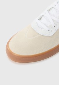Sneakers com parte superior em camurça bege clara, atacadores brancos e uma entressola texturizada, apresentando uma sola de borracha caramelizada para tração e aderência.