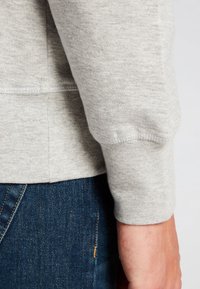 Sudadera gris con una tela suave y texturizada y un puño ajustado, combinada con vaqueros azul oscuro, mostrando detalles de costura en el dobladillo.