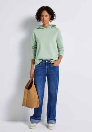 Haut à capuche vert clair avec cordon, jeans en denim bleu et sac fourre-tout en daim beige. Baskets blanches avec une semelle texturée. Tenue décontractée.