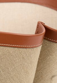 Gros plan d’un tissu beige tissé avec bordure en cuir marron et coutures blanches sur un bord incurvé.