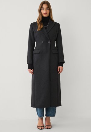Woman wearing a long dark gray double-breasted coat over a black turtleneck, cropped blue jeans, and black open-toe heels on a plain background.