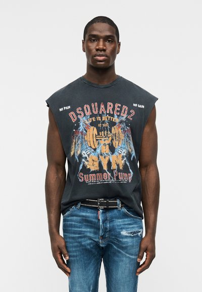 Άνδρας που φορά αμάνικο μαύρο γυμναστηριακό t-shirt με γραφικά και κείμενο, και μπλε τζιν, στέκεται μπροστά σε απλό ανοιχτό γκρι φόντο.