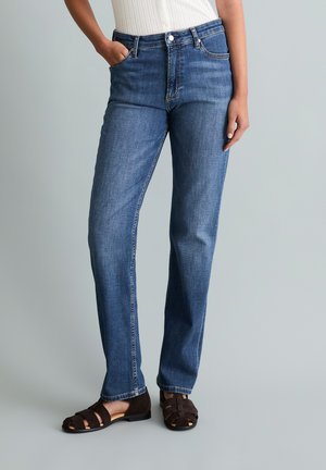 Person trägt gerade geschnittene blaue Jeans, weißes Oberteil und dunkelbraune geschlossene Sandalen, steht mit einer Hand in der Tasche.