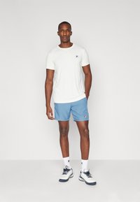 White short-sleeve shirt with blue logo, paired with blue shorts. Footwear is white with navy accents. Model stands in a neutral background.