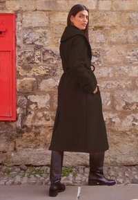 Palton lung negru cu guler lat și curea, realizat dintr-un material neted. Prezentă buzunare și are o croială dreaptă, completat de cizme negre până la genunchi.