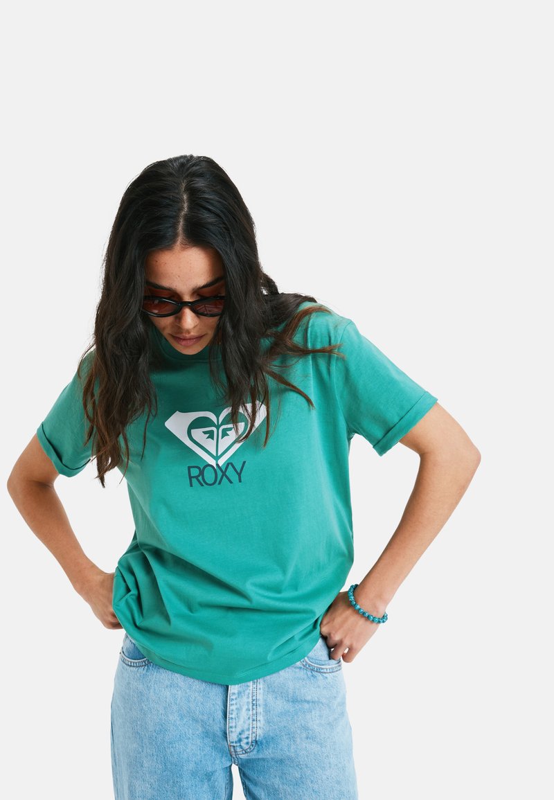 Tyrkis bomulds-t-shirt med et hvidt hjerte-logo og teksten "ROXY", parret med lyseblå denimjeans. Stoffet ser blødt og lidt afslappet ud.