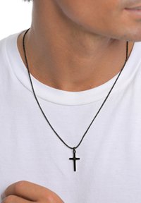 Schwarzes Kreuz-Anhänger an einer dünnen, geflochtenen schwarzen Kordelkette, getragen über einem weißen T-Shirt. Keine weiteren dekorativen Elemente.