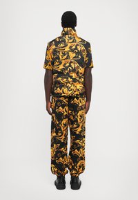 Homme de dos portant une chemise à manches courtes à motifs baroques noirs et dorés, un pantalon assorti et des chaussures à plateforme noires.