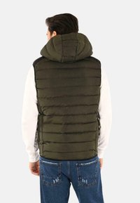 Gilet da puffer verde con cappuccio, design trapuntato, materiale leggero e zip frontale. Indossato sopra una maglietta bianca a maniche lunghe.