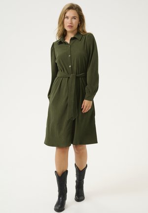 Robe en velours côtelé vert kaki avec un devant à boutons, un col, des manches longues, une ceinture à la taille et un tissu texturé, associée à des bottes de cowboy noires.
