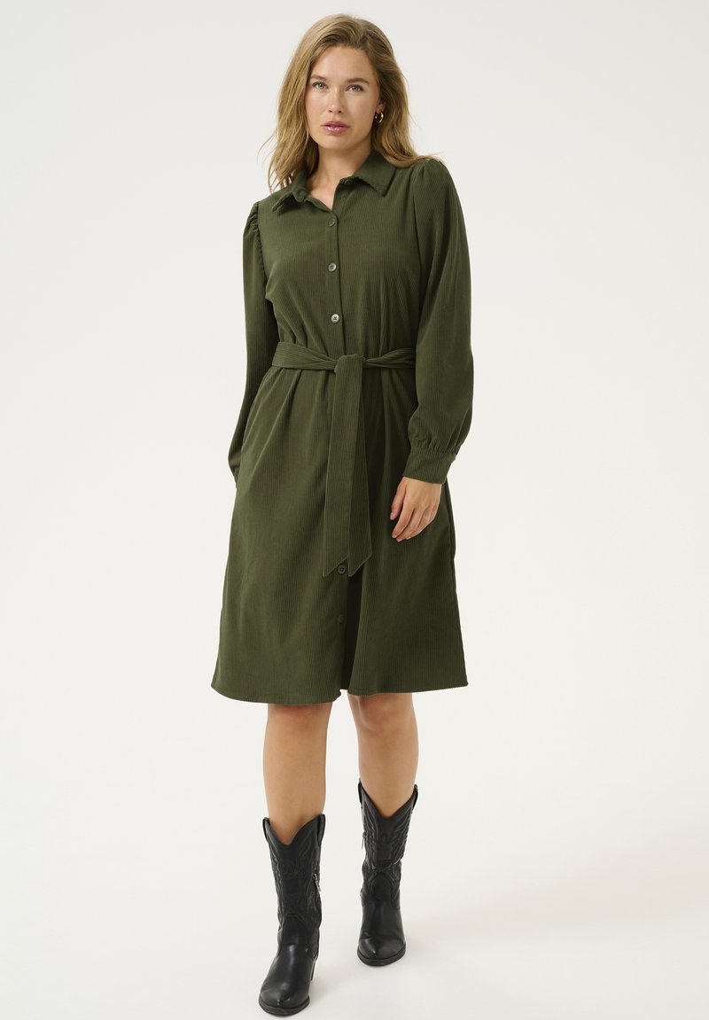 Robe en velours côtelé vert kaki avec un devant à boutons, un col, des manches longues, une ceinture à la taille et un tissu texturé, associée à des bottes de cowboy noires.