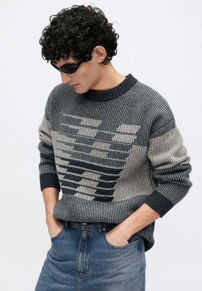 Camisola de malha em tons de cinza com um padrão geométrico, apresentando gola e punhos canelados, combinada com jeans azul denim.