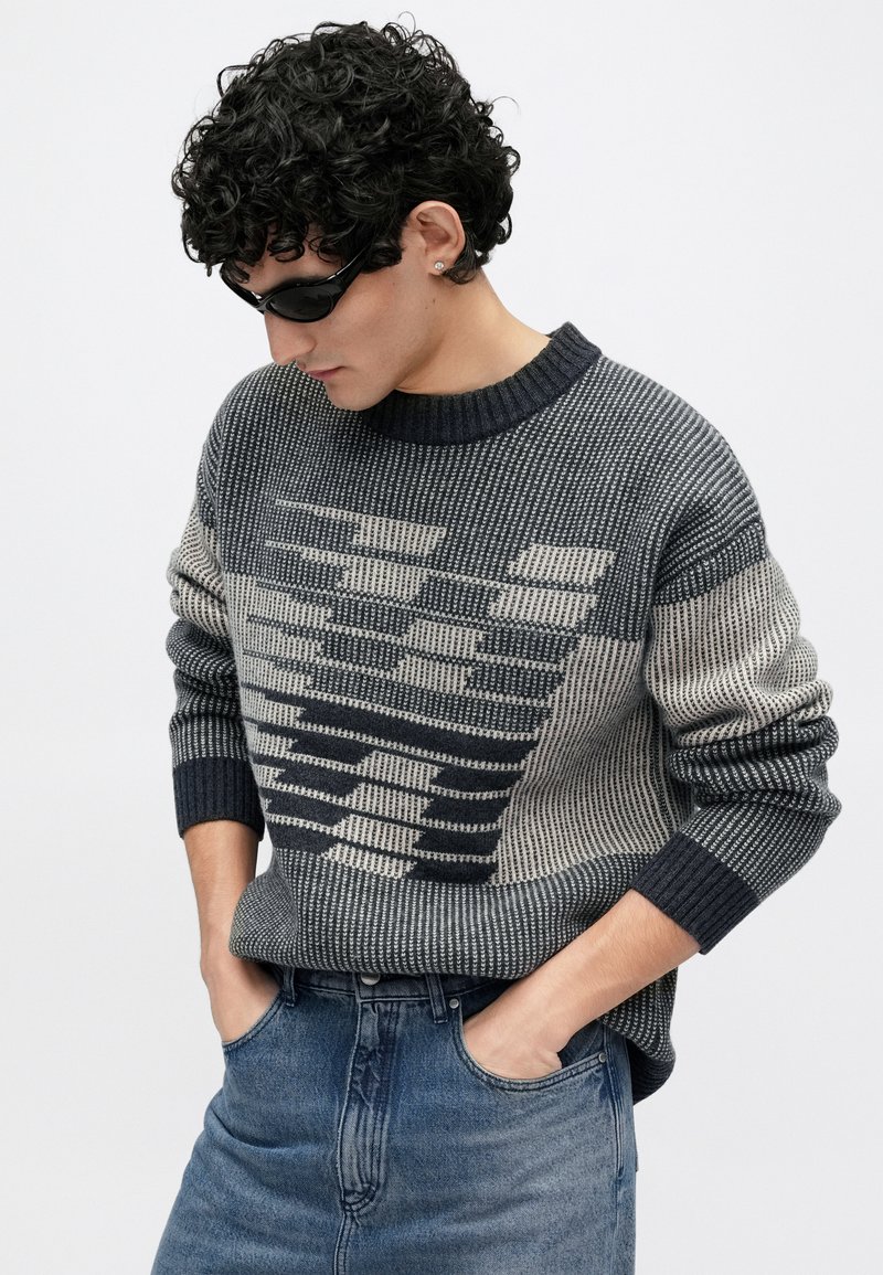 Pull tricoté dans des tons gris avec un motif géométrique, doté d'un col et de poignets côtelés, associé à un jean en denim bleu.