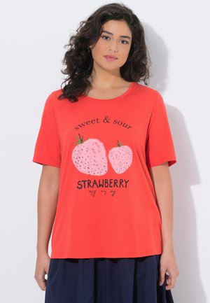 Kvinde med krøllet mørkt hår iført en rød T-shirt med illustrerede jordbær og teksten "sweet & sour STRAWBERRY enjoy every day."