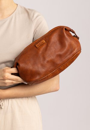 Žena v béžovém topu drží velkou hnědou koženou kabelku s malou vyraženou nášivkou vpředu.