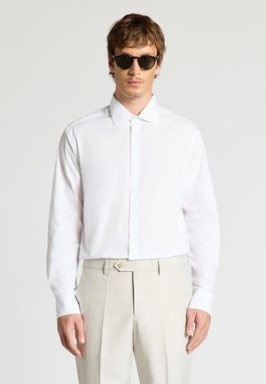 Homme portant des lunettes de soleil noires, une chemise blanche à manches longues boutonnée et un pantalon beige taille haute, debout devant un fond uni.