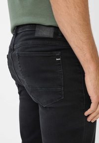 Schwarze Denim-Jeans im Slim-Fit-Stil mit einem Lederpatch mit der Aufschrift "Brax" am Bund und einem kleinen Logo auf der Gesäßtasche.