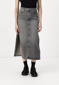 Denimrock in verblasstem Grau mit einem Schlitz vorne. Hat eine hohe Taille, Gürtelschlaufen, Vordertaschen und eine rohen Saum. Getragen mit schwarzen Sneakers.