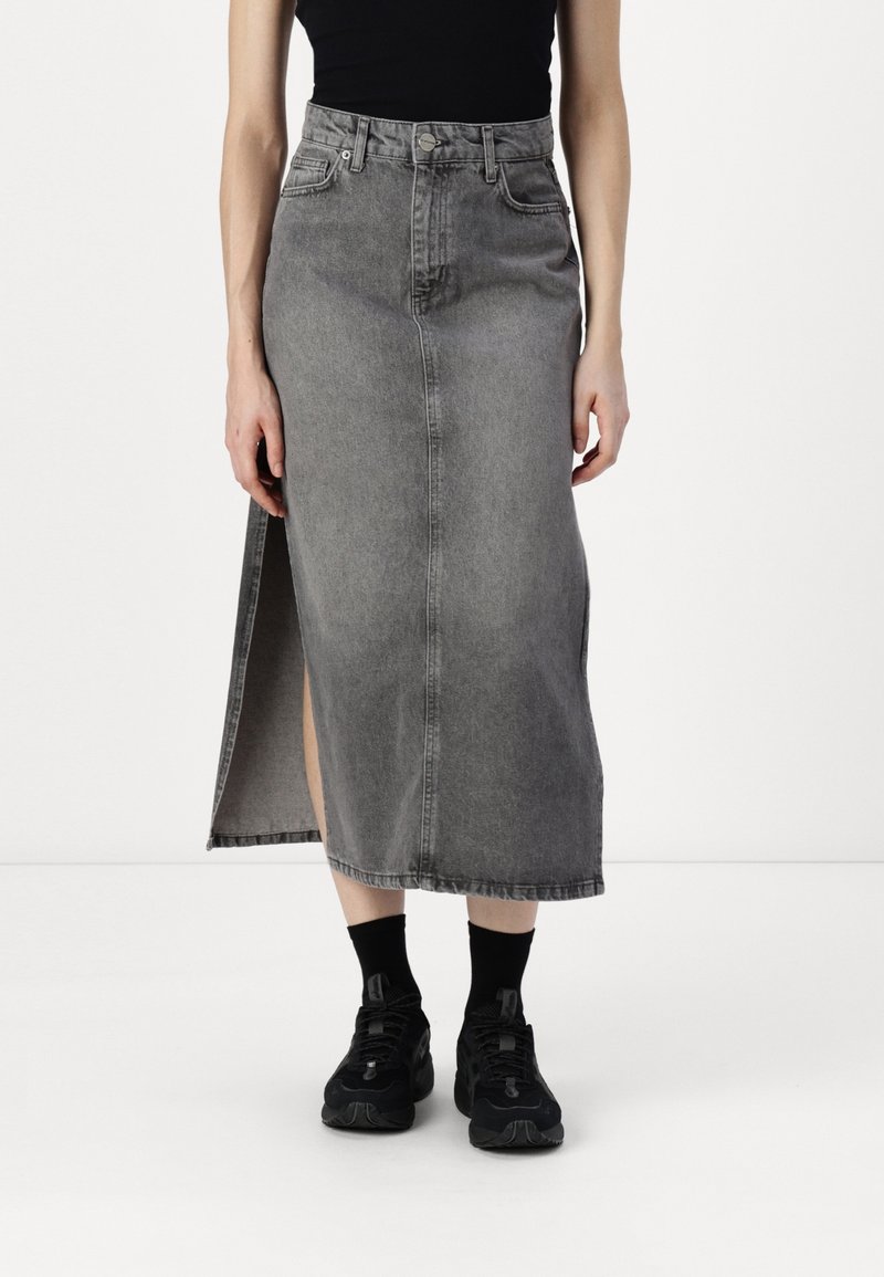 Denimrock in verblasstem Grau mit einem Schlitz vorne. Hat eine hohe Taille, Gürtelschlaufen, Vordertaschen und eine rohen Saum. Getragen mit schwarzen Sneakers.