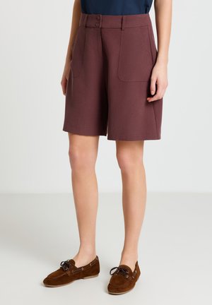 Femme portant un short marron mi-long avec poches avant et fermeture à double bouton, associé à des mocassins en daim marron à lacets.