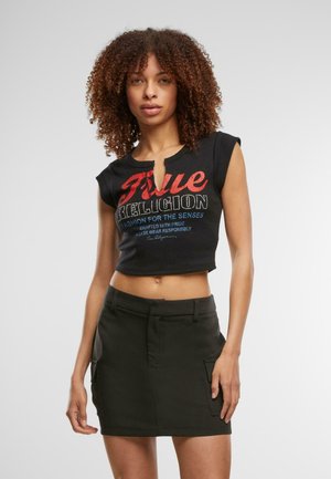Frau mit lockigem Haar, die ein schwarzes, kurzes T-Shirt mit rotem und blauem Text sowie einen schwarzen Minirock mit seitlichen Taschen vor einem einfarbigen Hintergrund trägt.