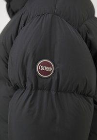 Fekete puffdzseki, amelyen egy kiemelkedő, bordó és ezüst Colmar logó jelvény található a bal karon, sima, texturált felszínggel.