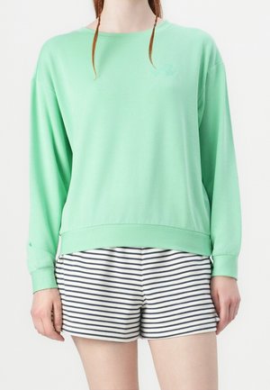 Personne portant un haut à manches longues vert menthe et un short blanc à rayures horizontales noires sur un fond neutre.