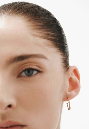 Pendiente de aro dorado con forma de imperdible, colgando del lóbulo de la oreja, con una superficie texturada y un brillo sutil sobre un fondo neutro.