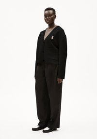 Svart kort cardigan med V-ringning, enda rosa banddetalj, som är ihop med svarta vida byxor och tvåfärgade skor. Slät textur.