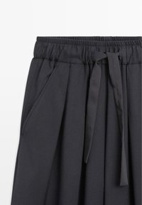 Black, elastic waistband skirt with pleats, featuring a tie detail and side pockets. Made from a smooth fabric with a matte finish.