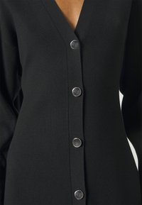 Cardigan noir en tricot avec un col en V profond et cinq boutons ronds argentés sur le devant. Le tissu présente une texture côtelée.