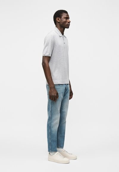 Άνδρας σε προφίλ που φοράει ανοιχτό γκρι κοντομάνικη πόλο, μπλε τζιν, λευκές κάλτσες και λευκά αθλητικά παπούτσια με φόντο απλό.