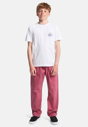 T-shirt blanc en coton avec un petit motif bleu sur la poitrine, associé à un pantalon rose ample et des baskets noires.