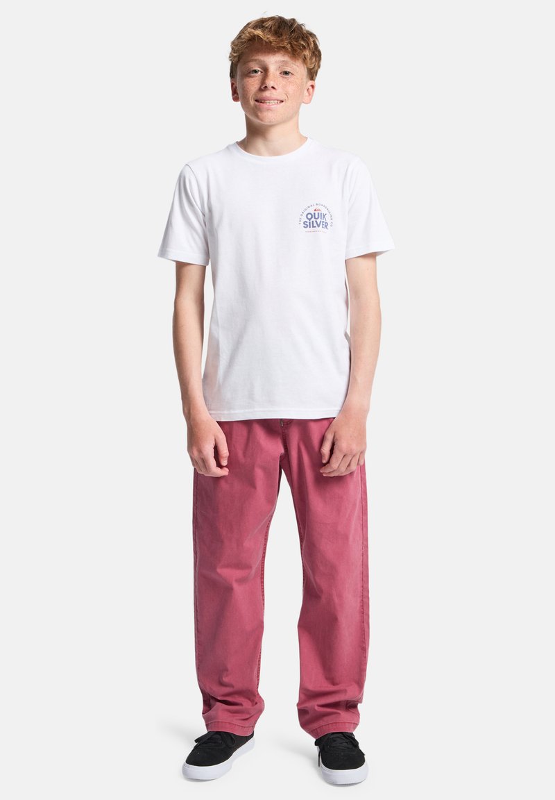 T-shirt blanc en coton avec un petit motif bleu sur la poitrine, associé à un pantalon rose ample et des baskets noires.