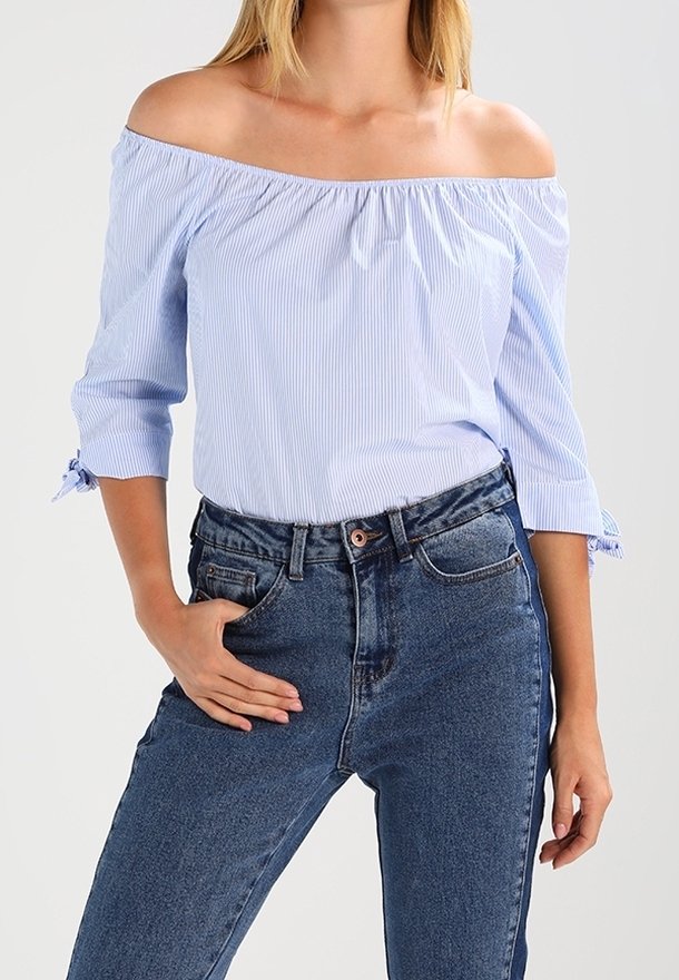 Femme portant un chemisier bleu clair à épaules dénudées avec des manches trois-quarts et un jean bleu taille haute, main dans la poche.