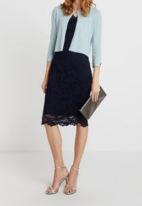 Himmelblauer, kurz geschnittener Cardigan über einem dunkelblauen Spitzenkleid mit gewelltem Saum, kombiniert mit silbernen Riemchenhemdchen und einer metallischen Clutch.