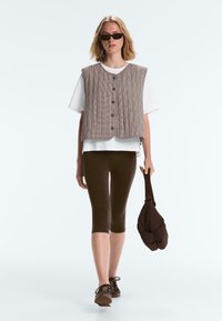 Femme portant des lunettes de soleil, un gilet matelassé marron sur un T-shirt blanc, des leggings capri marron foncé, des chaussures marron, tenant un petit sac marron.