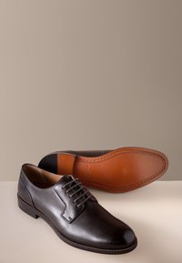 Paar polierte schwarze Lederschuhe, eines aufrecht stehend, das andere zeigt die glatte hellbraune Ledersohle mit genähten Kanten.