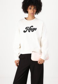 Valkoinen collegepusero, jossa on musta "Hugo"-logo, rento istuvuus. Mustat housut, käsissä vaaleanpunainen pikkulaukku, jossa on vetoketju ja logo-yksityiskohta.