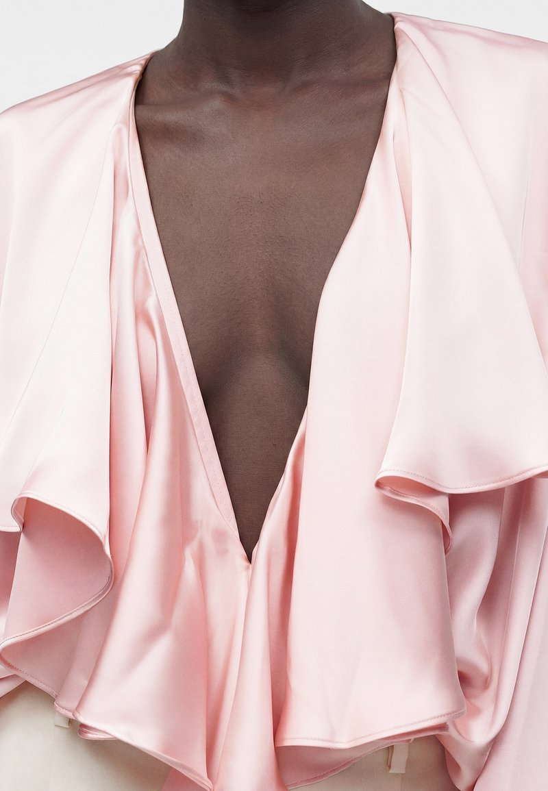 Personne à la peau foncée vêtue d'un chemisier en soie rose clair avec un col en V profond et un col à volants visible des épaules jusqu'à la poitrine.