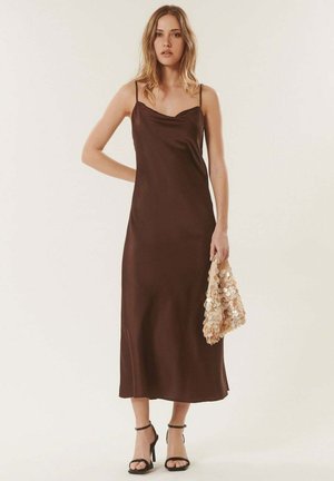 Jeune femme portant une robe nuisette marron et des talons noirs, tenant un sac à main beige à sequins sur un fond clair uni.