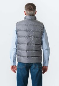 Grå polstret vest med høj krave, med horisontale quiltede sektioner. Bæres over en lyseblå stribet skjorte og et par denim jeans.