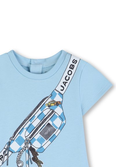 Jasnoniebieska koszulka z krótkim rękawem, na której znajduje się grafika w postaci niebieskiej kratkowanej saszetki biodrowej, żółtego taksówki oraz czarnego logo na pasku.