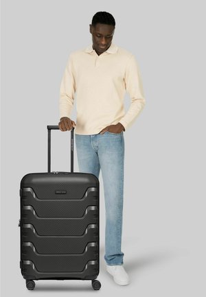 Man in beige long-sleeve shirt and jeans standing next to a black hard-shell suitcase with extended handle on gray background.